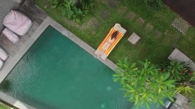Havadan bej bikinili genç bir kadının rahatlama görüntüsü ve Ubud 'daki şezlonglu ormanda yüzme havuzunun yanında güneş banyosu yapması. Drone yükseliyor. Tatil kavramı.