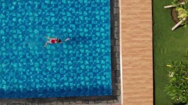 Kırmızı bikinili bir kadın havuzda kurbağalama gibi yüzüyor ve havuzdan çıkıyor. Sportif bayan yüzücü. Hava manzaralı. İHA