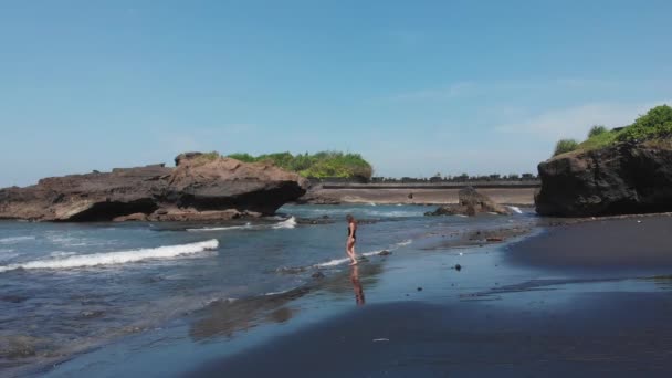 Aerial drone view of woman with sexy butt in black swimsuit go into the ocean with foam waves on black sand beach with many stones and rocks in ocean. Bali Island, Indonesia. — Wideo stockowe