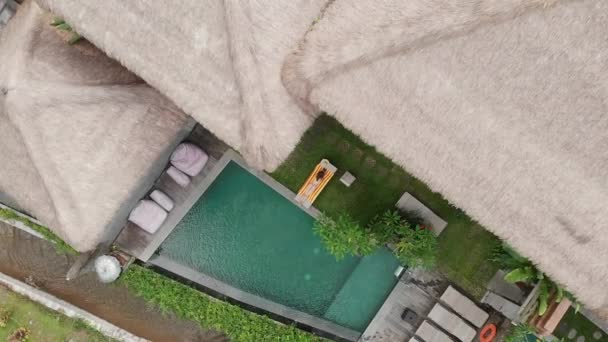 Vista aérea de la villa con techos de paja con piscina y mujer joven delgada en bikini beige relajante y tomar el sol en chaise-longue en el jardín tropical Ubud. Drone cayendo. Concepto de vacaciones . — Vídeos de Stock