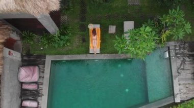Hava aracı, bej bikinili, zayıf genç bir kadının Ubud 'daki şezlonglu ormanda havuz kenarında güneşlenişini gösteriyor. Tatil kavramı.