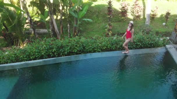 Luftaufnahme des Infinity-Pools auf einer exotischen Luxusinsel. Frau, die am Rande des Pools spaziert und in rotem Bikini und Hut den Blick auf den Dschungel genießt. Urlaubskonzept — Stockvideo