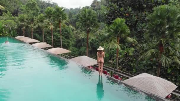 Aerial Top Down View of Infinity pool at luxurious exotic island. Back view of woman walking on edge of pool and enjoy jungle view wearing beige bikini and hat. Vacation concept — Stock Video