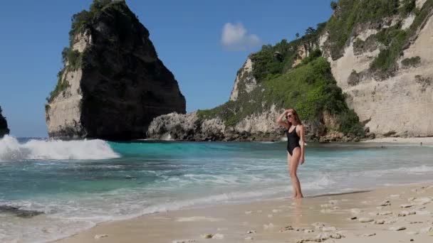 Hezká žena se šťastně prochází a užívá si podél pobřeží oceánu s kameny a tyrkysovým oceánem, modrá obloha. Pláž Atuh, ostrov Nusa Penida, Bali, Indonésie. Tropická koncepce cestování. — Stock video