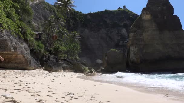 Mooie vrouw loopt naar de oceaan met zee rotsen en turquoise oceaan, blauwe lucht. Strand van Atuh, Nusa Penida eiland, Bali, Indonesië. Tropische achtergrond en reisconcept. — Stockvideo