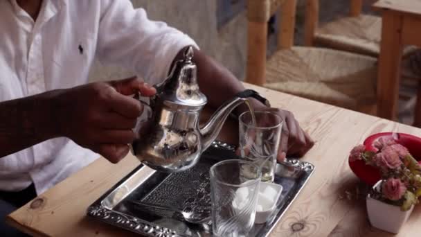 Mão masculina derramando chá de bule de prata ornamentado em copos em pequena mesa de acordo com a tradição marroquina. Preparação ritual de chá de hortelã em Marrocos — Vídeo de Stock