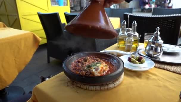 Tradicional marroquí comida tajina cocinar en macetas en la mesa en la cafetería y el gato disfrutando de la vista. Mano del cocinero local está abriendo plato hirviendo y liberar humo en tiempo soleado brillante — Vídeo de stock