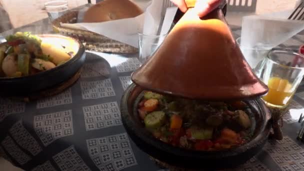 Tradicional marroquí comida tajina cocinar en macetas en la mesa en la cafetería con verduras en la parte superior. Mano del cocinero local está abriendo plato hirviendo y liberar humo en tiempo soleado brillante — Vídeo de stock