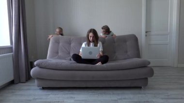 Modern evde oynayan kızları dikkatini dağıtırken annesi laptopta evden kanepeye oturuyor. Kendini soyutlama. Karantina sırasında sosyal uzaklık