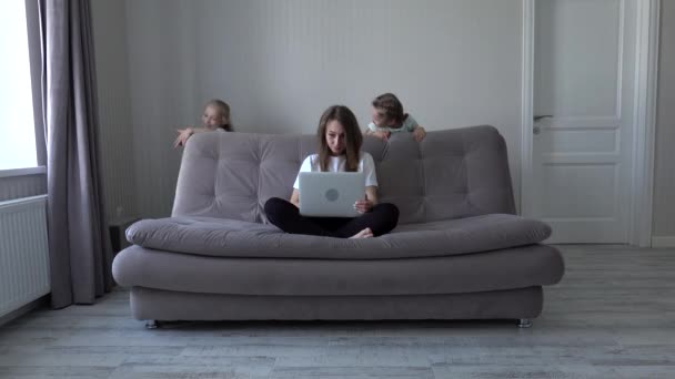 Мать, работающая за ноутбуком из дома, сидит на диване, в то время как ее дочери, играющие в современном доме, отвлекают ее. Самоизоляция. социальное расстояние во время карантина — стоковое видео