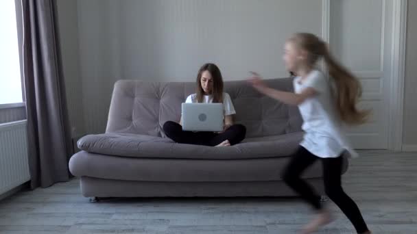 Madre che lavora al computer portatile da casa seduta sul divano mentre le sue figlie che corrono a giocare in casa moderna la distrae. Auto-isolamento. distanza sociale durante la quarantena — Video Stock