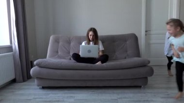 Modern evde koşuşturan kızları dikkatini dağıtırken, dizüstü bilgisayar başında çalışan kadın anne kanepeye oturuyor. Kendini soyutlama. Karantina sırasında sosyal uzaklık