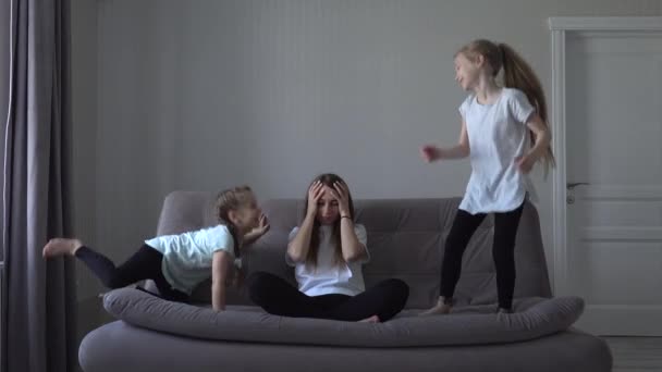 Estressado exausto chateado mãe sentindo dor de cabeça cansado de crianças ativas, jovem mãe sentar no sofá se sentir frustrado com as filhas barulhentas correndo brincando em casa moderna . — Vídeo de Stock