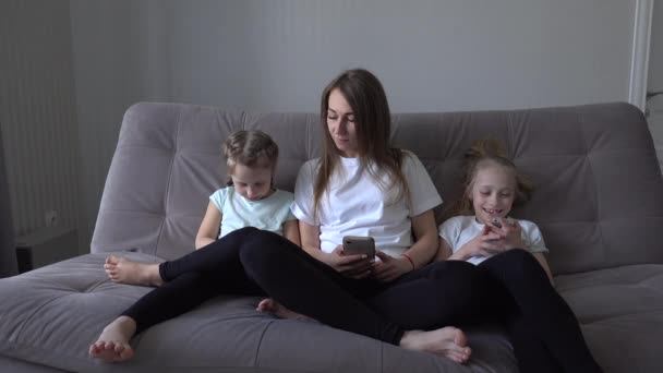 A boldog család portréja, amint a szürke kanapén ülnek személyes holmikkal. Anya és gyermekei otthon töltik az idejüket a modern technológiával.. — Stock videók