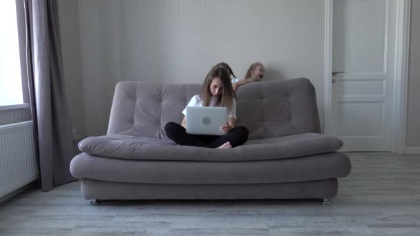 Семья на карантине. Уставшая мать пытается работать из дома на ноутбуке, сидя на диване под напряжением двух шумных детей, бегающих вокруг. Самоизоляция. Социальное расстояние во время карантина — стоковое видео