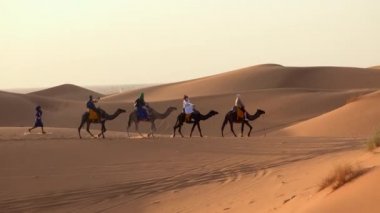 Sahra çölünde deve kervanı gün batımında ve mavi gökyüzü arka planında, Fas 'ta. Turistler kum tepelerinde deveye binerler. Çöl konvoyunda deve sahipleri