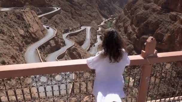 Widok z tyłu turystki w białej sukience podnoszącej ręce i cieszącej się widokiem na wietrzną górską drogę w wąwozie Dades, Gorges Du Dades, Maroko — Wideo stockowe