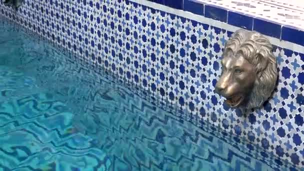 MARRAKESH, MOROCCO - OUTUBRO 2019: Vista de perto da piscina com mosaico azulejo cabeça azul e branca e dourada de leão em design tradicional árabe em riad marroquino . — Vídeo de Stock