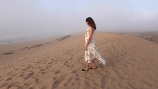 Jovem mulher caminhando ao longo da praia de areia do mar em dia de nevoeiro chuvoso. Menina em vestido longo desfrutar de vista sobre duna de areia perto do oceano em Marrocos — Vídeo de Stock