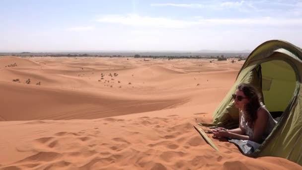 Туристка, лежащая в палатке в пустыне Сахара среди песчаных дюн и наслаждающаяся диким ландшафтом в Марокко — стоковое видео