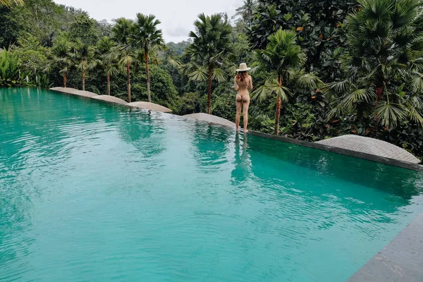 Infinity pool at luxurious exotic island. Back view of woman walking on edge of pool and enjoy jungle view wearing beige bikini and hat. Vacation concept.