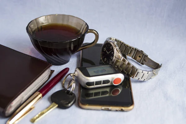 Bir fincan kahve, bir cep telefonu, bir defter, tükenmez kalem, arka planda anahtarlık olan bir araba anahtarı, sabah.