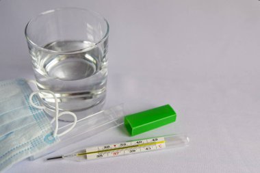 Bir bardak su, yüksek sıcaklık göstergesi olan bir termometre, tıbbi maske, haplar, akan bir burun için damla. Beyaz arka plan, kimse yok.