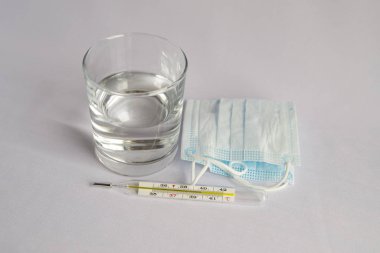 A glass of water, a thermometer with a high temperature, a medical mask. White background, no one.