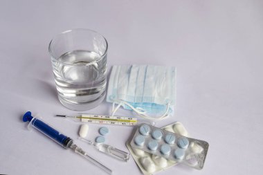 Bir bardak su, yüksek sıcaklık göstergesi yakın bir termometre, tıbbi maske, haplar, tıbbi şırınga. Beyaz arka plan, kimse yok.
