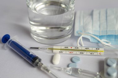 Bir bardak su, yüksek sıcaklık göstergesi yakın bir termometre, tıbbi maske, haplar, tıbbi şırınga. Beyaz arka plan, kimse yok.