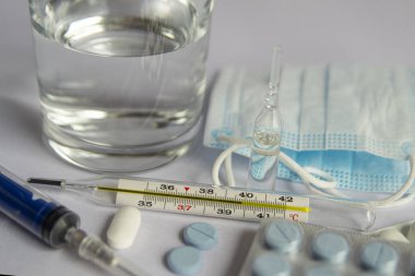 Bir bardak su, yüksek sıcaklık göstergesi yakın bir termometre, tıbbi maske, haplar, tıbbi şırınga. Beyaz arka plan, kimse yok.