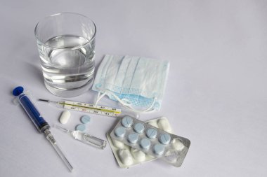 Bir bardak su, yüksek sıcaklık göstergesi yakın bir termometre, tıbbi maske, haplar, tıbbi şırınga. Beyaz arka plan, kimse yok.