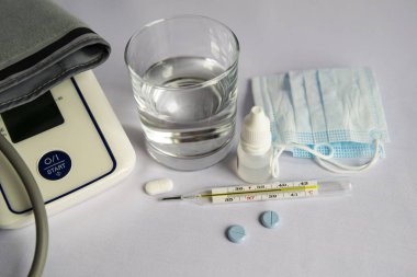 Bir bardak su, yüksek sıcaklık göstergesi olan bir termometre, tıbbi maske, haplar, akan bir burun için damla. Beyaz arka plan, kimse yok.