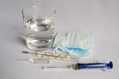 Bir bardak su, yüksek sıcaklık göstergesi yakın bir termometre, tıbbi maske, haplar, tıbbi şırınga. Beyaz arka plan, kimse yok.