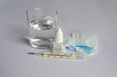 Bir bardak su, yüksek sıcaklık göstergesi yakın bir termometre, tıbbi maske, haplar, tıbbi şırınga. Beyaz arka plan, kimse yok.