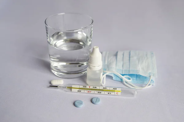 Bir bardak su, yüksek sıcaklık göstergesi yakın bir termometre, tıbbi maske, haplar, tıbbi şırınga. Beyaz arka plan, kimse yok.