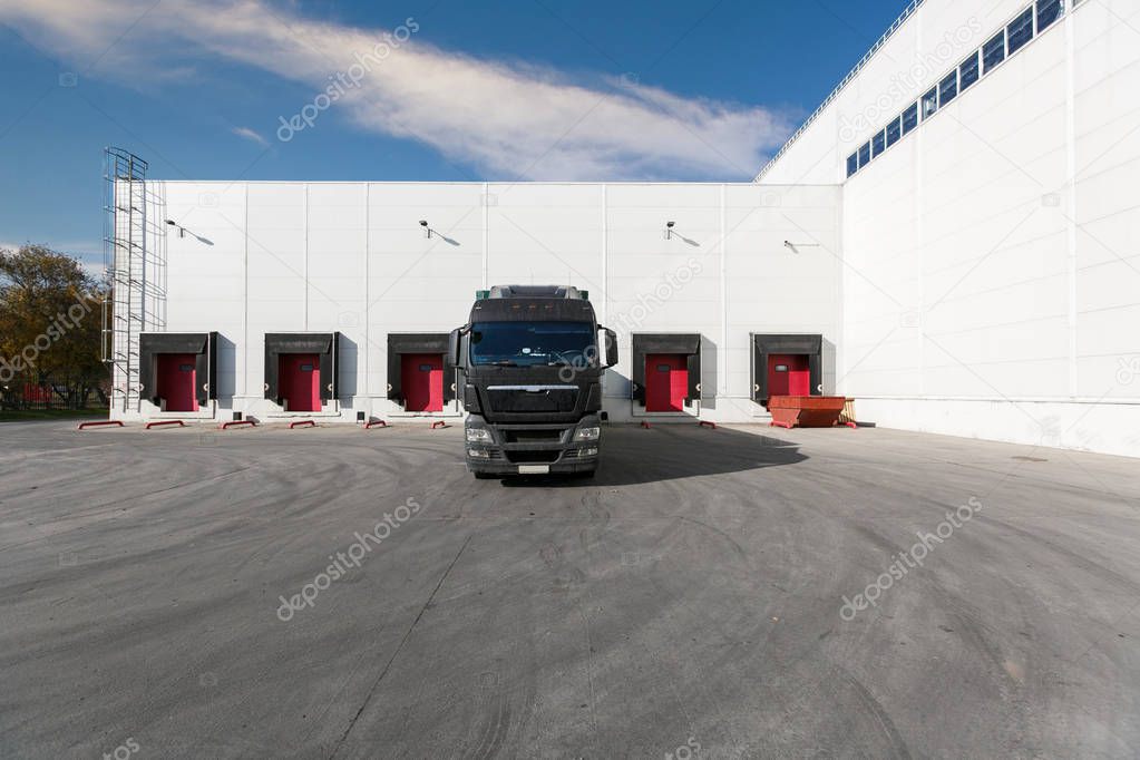 black truck with a blue trailer at unloading at the warehouse.Warehouse complex with an asphalt pad and truck with blue sky
