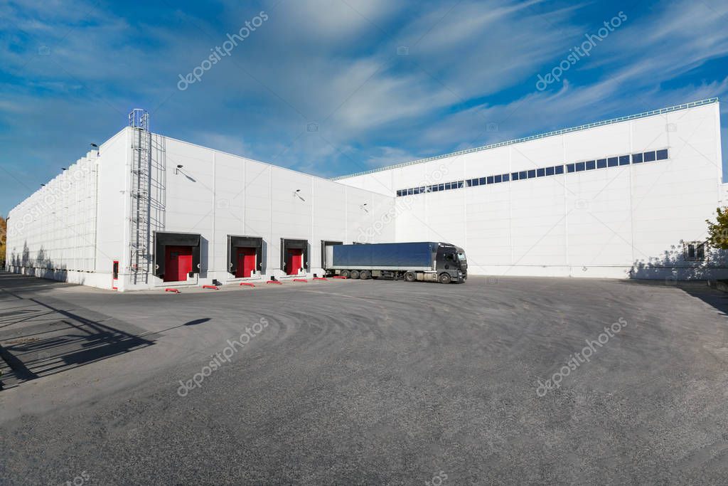black truck with a blue trailer at unloading at the warehouse.Warehouse complex with an asphalt pad and truck with blue sky