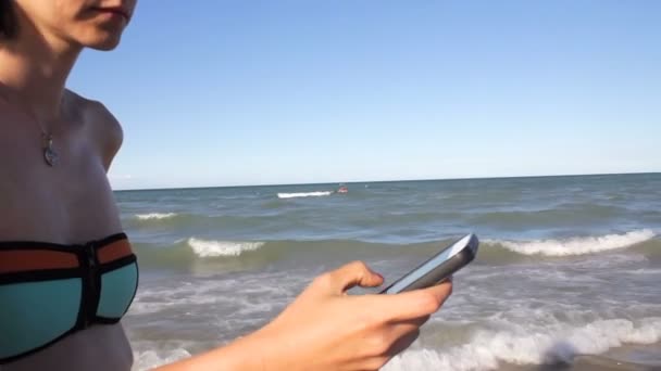 Mujer en bikini SMS mensajes de texto utilizando la aplicación en el teléfono inteligente en la playa. Primer plano de las manos utilizando el teléfono inteligente al aire libre. Teléfono celular móvil de cerca . — Vídeos de Stock