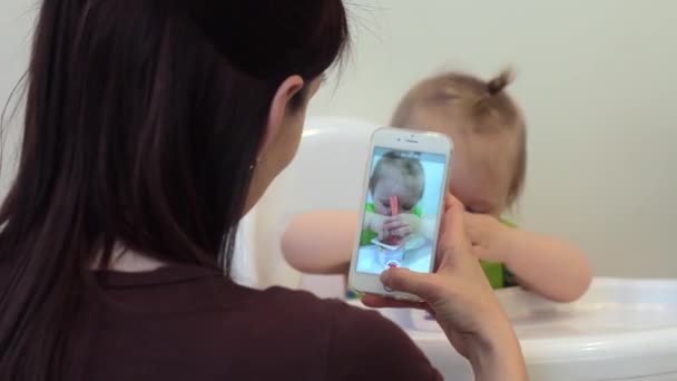 Mamma tar foto och video av barnet med hjälp av smartphone njuter av att fotografera söta barn dela moderskap livsstil på sociala medier. Flickan äter för första gången — Stockvideo