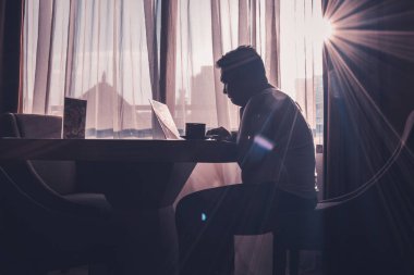 The silhouette of a man at work using a laptop in a modern room with a window background illuminated by slightly dazzling natural sun glare.