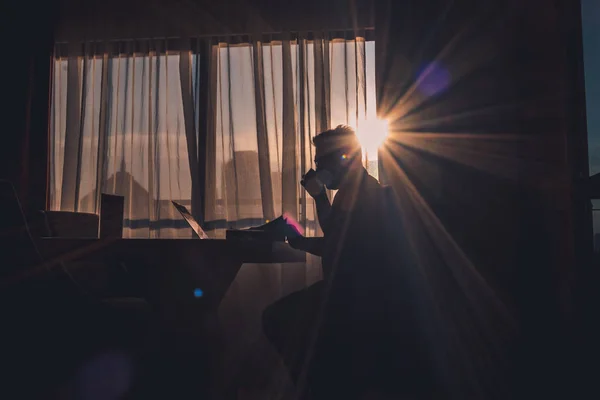 The silhouette of a man drinking coffee while working on a laptop in a modern bedroom with a window background lit by dazzling sunlight.