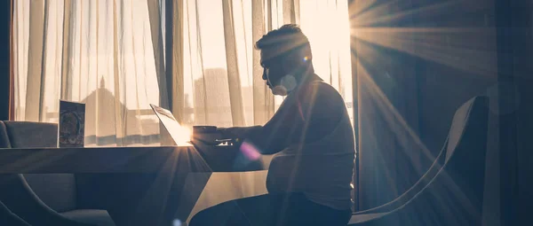 The silhouette of a man at work using a laptop in a modern room with a window background illuminated by slightly dazzling natural sun glare.
