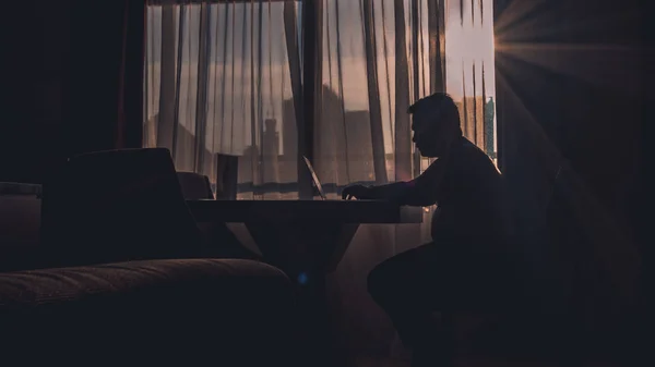 The silhouette of a man at work using a laptop in a modern room with a window background illuminated by slightly dazzling natural sun glare.