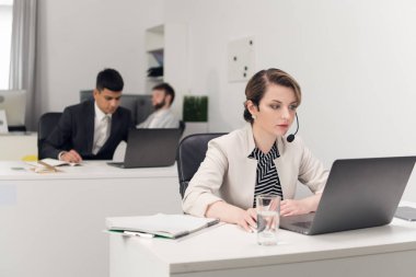 Mikro bilgisayarı olan bir kadın dizüstü bilgisayarla masada oturuyor, meslektaşlar işleriyle meşgul.