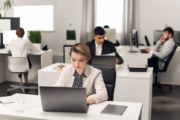 Frau arbeitet am Laptop, Männer hinter ihr blättern in Bürounterlagen — Stockfoto