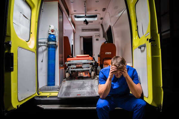 Запутавшийся санитар в синей форме сидит на машине скорой помощи.
