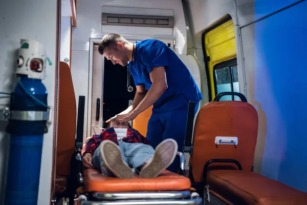 Мужчина в медицинской форме надевает кислородную маску на бесчувственную женщину, лежащую на носилках в машине скорой помощи — стоковое фото
