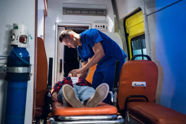 Парамедик в форме надевает кислородную маску на бесчувственную женщину, лежащую на носилках в машине скорой помощи. — стоковое фото