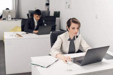 Takım elbiseli insanlar ofiste dizüstü bilgisayarlar ve mikrofonlarla çalışıyorlar.